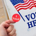 Photograph of a person holding an "I Voted" sticker in front of a "Vote Here" sign with an American Flag on it.